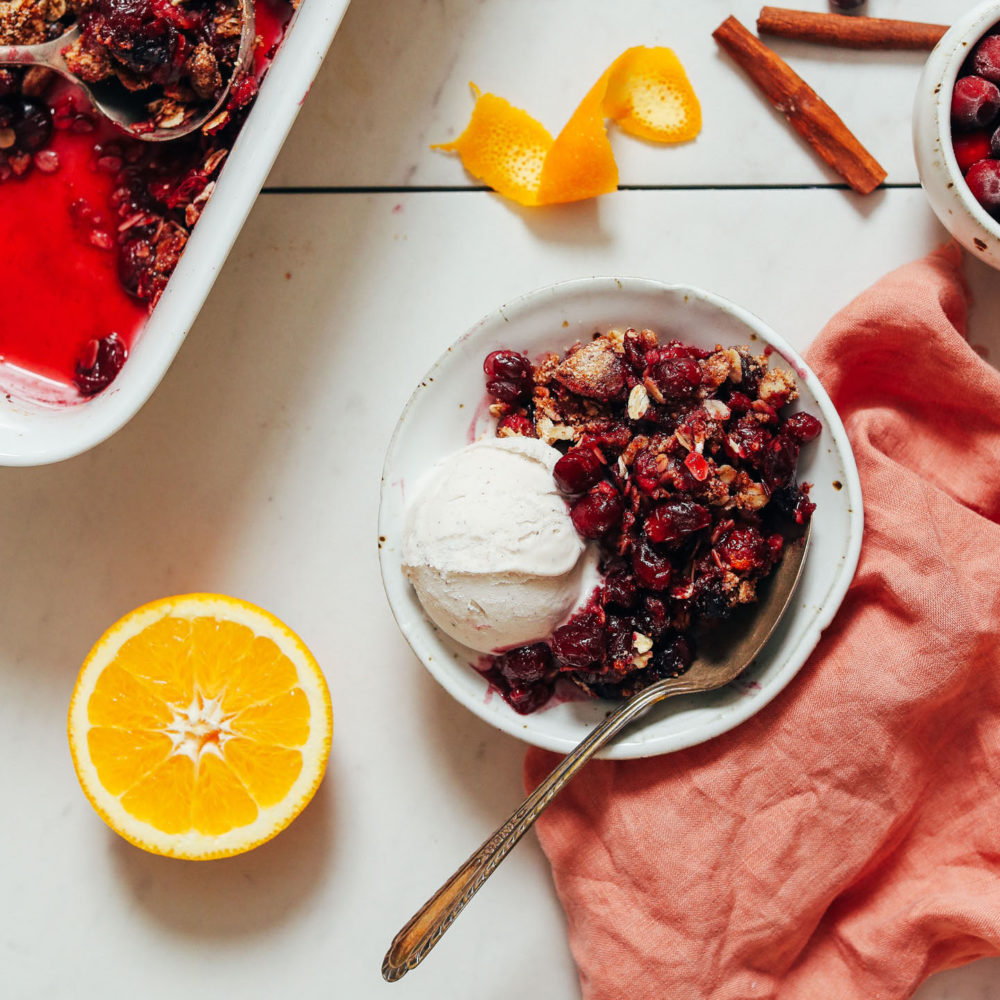 Sliced orange and cinnamon sticks next to a bowl and baking dish of orange cranberry crisp