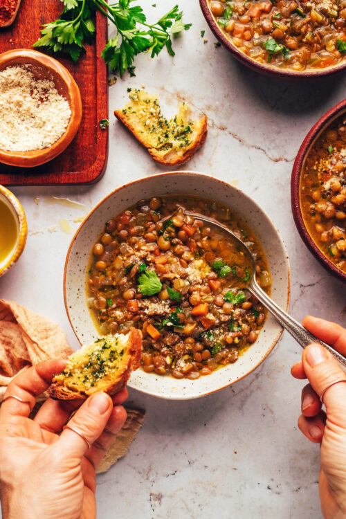 Holding a slice of garlic bread in one hand and a spoon in the other in a bowl of Italian lentil soup